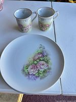 Two mugs and one plate showing floral pattern and gold rim