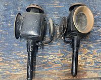 pair of black metal carriage lamps lying side by side showing front circular lenses and mounting posts