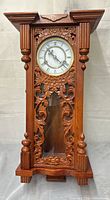 Front view of carved wooden pendulum wall clock showing dial, carvings, glass panel and pendulum