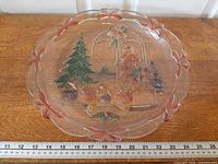 Full top view of clear glass serving platter showing scalloped rim, bow accents and central painted holiday scene beside measuring scale