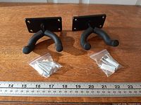 Two black wall-mount guitar hangers with foam-coated forks and mounting hardware packets on wooden surface with ruler for scale