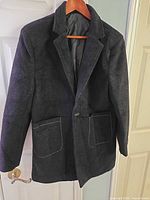Front view of black blazer on hanger showing snap closure and patch pockets with stud trim