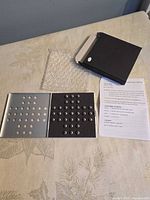 Open hinged aluminum board showing peg holes and marbles beside box and instructions