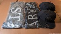 Three scarves in retail plastic and three heather gray caps stacked