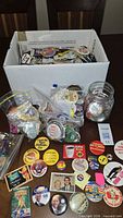Open box showing large quantity of assorted pinback buttons, some in bags and jar