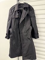 Front view of dark double-breasted trench coat on hanger showing buttons, lapels, belt and cuff straps