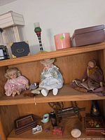 Overall view of three shelves showing lamp shade, boxes, candle holder, planter, dolls, figurines, vase, jewelry box and other small items