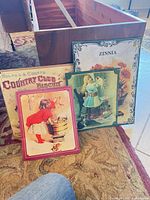 Three reproduction tin signs leaning against chest: Country Club Biscuit, Zinnia, girl at washtub