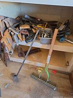 Shelf with pile of hand saws, chisels, bin of small tools, C-clamp, crow bar, two yard tools in foreground