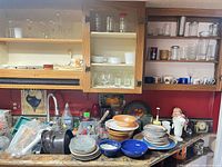 Wide view of countertop and upper cabinets showing stacked ceramic plates and bowls, clear glass bowls, assorted glasses and mugs, metal jug, utensils and cleaning items