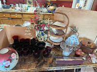 Overview of lot on countertop showing red glassware, wicker stand with plates, vase with flowers, casserole dish, pear container and boxed chopsticks