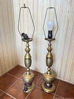 Two brass table lamps standing side by side on tile floor, showing full bodies, harps, cords, one bulb present