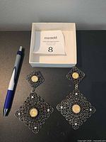 Overall view showing both earrings, detailing filigree pattern and yellow cabochons, pen for scale