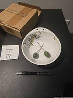 Top view of ceramic dish with olive motif, two picks inside, box and scale pen visible