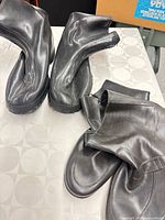 Two pairs of black footwear: leather boots and rubber overshoes displayed on table