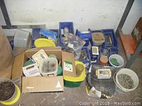 Photo showing various bins, buckets, cardboard boxes, and plastic containers filled with different screws and nails scattered on the floor.