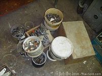 Overview of five plastic buckets containing assorted nuts, bolts, hooks, wrenches, and hardware items on floor.