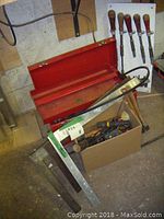 Photo showing red metal toolbox, cardboard box of assorted hand tools, metal square, and a large metal tool with wooden handle.
