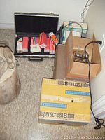 Photo showing the open cardboard box labeled 'Superscreen 8mm' with projector inside and a black briefcase on the floor containing projector lamp boxes.