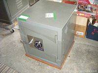 Gray Sears floor safe in a room with concrete floor, metal industrial shelving nearby and assorted boxes. The safe is a cubic form with a metal door featuring combination dial and handle.