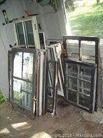 Group of eight storm windows leaning against a wall showing varied sizes and damaged frames.