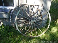 Two antique wooden wagon wheels leaning against a building on grass, showing weathered green paint and rusted metal parts.