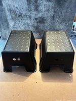 Two black IKEA BOLMEN plastic step stools side by side, upside down showing undersides