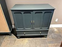 Front view of dark teal IKEA LOMMARP cabinet showing two doors, lower drawer, and brass pulls