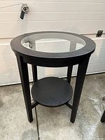 Front angled view of round black wood end table with glass inset top and lower shelf