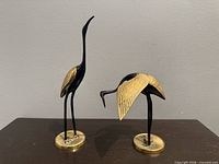 Both brass and black enamel crane figurines, upright and bending, on dark surface