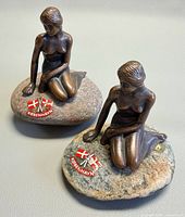 Front angled view of both mermaid sculptures on stone bases showing decals and patina