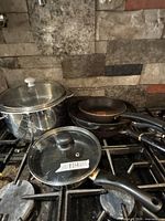 Cookware set on stovetop: stainless stock pot with lid, two nested nonstick frying pans, glass lid labelled 114, saucepan handle visible