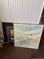 Both framed prints leaning against wall: swan print front, landscape behind