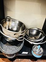Stacked stainless colanders, nested mixing bowls, black speckled roaster with lid, plastic salad spinner visible together