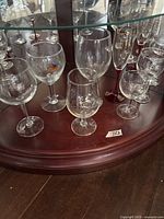 Group of assorted stemware on round wooden shelf showing Leffe goblet, wine glasses, red-stem flute and small goblets