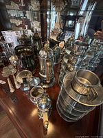 assortment of silver-plated coffee service pieces, condiment pots, servers and stacked stainless cups on display shelf