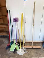 Long-handled brooms, mop, duster, can crusher, push broom, plunger, loose tool heads against wall