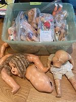 Plastic bin containing multiple bagged dolls and two large doll bodies displayed in front