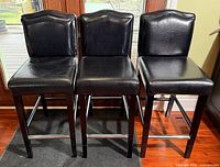 Front view of three identical black faux-leather counter stools on black wood frames with chrome footrests