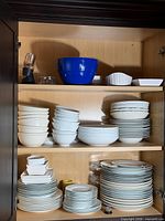 Cabinet shelves filled with stacked white plates, bowls, blue mixing bowl, electric grinder, baking dishes