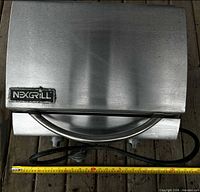 Top view of closed stainless Nexgrill portable grill with handle and hose visible, measuring tape shows scale
