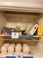 Shelf with boxed Chi Lava curling iron, metal sailboat statue, black hole punch, Sunbeam water bottle box, yellow first aid kit