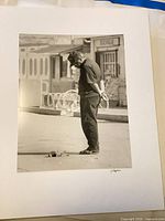 Matted B&W photo of man standing over small object, signature visible