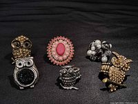 Five rings arranged on black fabric: three owl designs, pink floral, black & white cluster