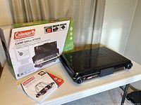 Boxed Coleman camp grill/stove with stove unit and manual on table