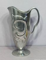 Full left-side view of aluminum pitcher displaying embossed scroll and pedestal base