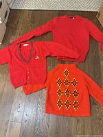 Three sweaters laid out: red crewneck, red cardigan, orange geometric pullover