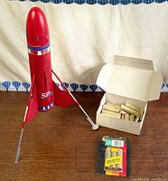 Overall shot of red rocket standing with landing legs, open box of rocket motors, sealed blister pack
