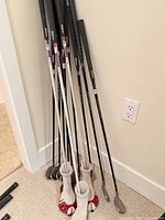 Group of right-handed golf clubs with graphite shafts leaning against wall; grips visible; four headcovers on woods