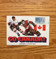 Front of postcard depicting players, stamp, flag, GO CANADA text